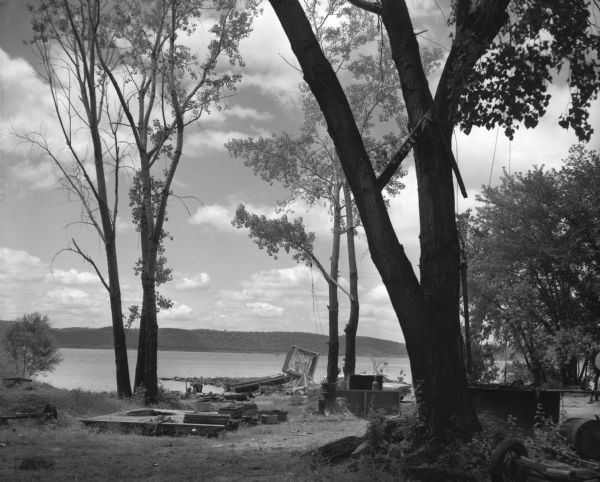A fishing dock along the shore of the Mississippi River. A man is in a boat near the shoreline, and an automobile is behind a tree. A number of boats are lined up along the bank along with a fish net drying reel.