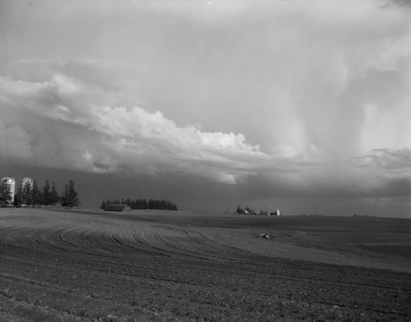 An upland field prepared for planting. In the distance, several farm buildings are visible beneath large storm clouds.
