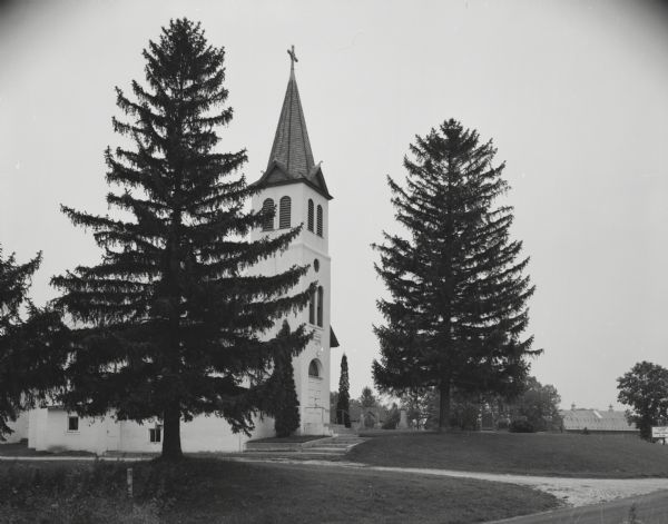 Exterior view of Saint Peter's Lutheran church. Set in a rural landscape, the church is flanked by two large pine trees. In the background on the right is the roof a large dairy barn.