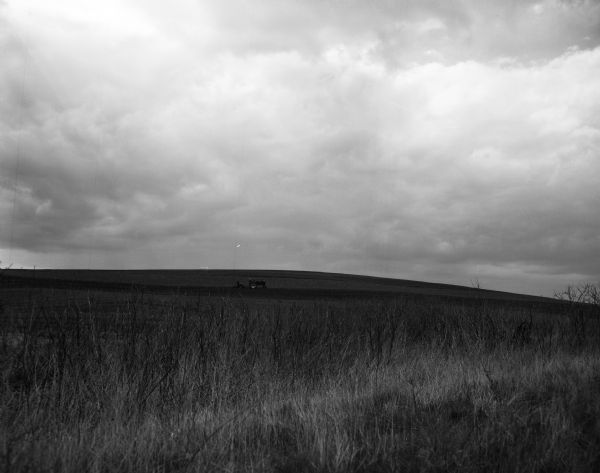 View uphill towards a farmer planting crops on a treeless slope which rises just beyond him, with a cloudy sky above. Various grasses are growing in the foreground.