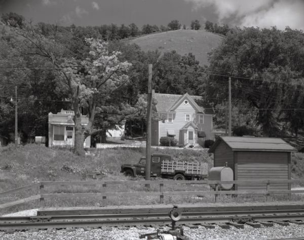View from the Mississippi River toward the bluffs. A residential home, old bank building, truck and railroad building are on a hill among trees below the bluffs. Railroad tracks run across the foreground.