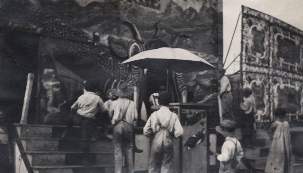 Children gathered in front steps and on a platform with large painted banners at a street carnival, possibly the State Fair. There is also a stand with an umbrella that is probably for selling tickets.