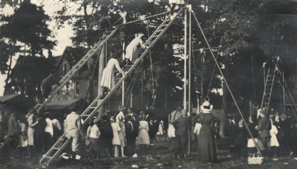 Children playing on the playground at the State Fair. The children form lines to climb up the two tall ladders to slide down a pole. Several children are near the top. There are also 'flying rings' on the right. Trees and buildings are in the background behind the ladder structure.