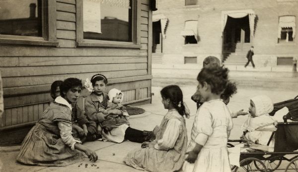 A group of girls playing jacks on a city sidewalk. All the girls are wearing dresses, and two infants are present, one in a carriage and one in the lap of a girl. A man is walking on the sidewalk across the street.