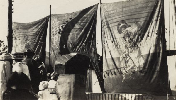 A group of adults and children are gathered in front of carnival banners. Probably depicting "Gay Paree" at the State Fair.