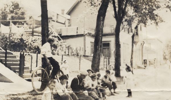 View from street of a group of ten children sitting on the curb of a street. Behind them are a house, trees, and a man on a bicycle. Seven of the ten children are boys, and almost all of them wear hats.