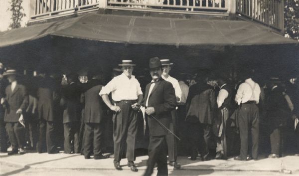 Three men wearing hats are standing in front of the bar at a Milwaukee picnic. Behind them are a large group of men wearing black suits crowded around the bar which is under an awning.