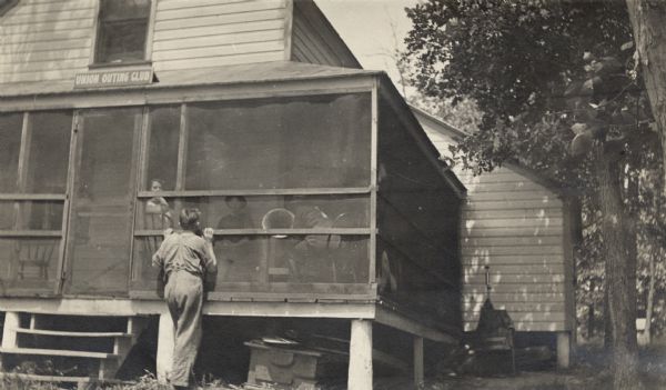 View from yard of a man standing at the rear of a cottage, with a screened-in porch that has a sign that reads "Union Outing Club" above the screen door. Several people are seated inside the porch.