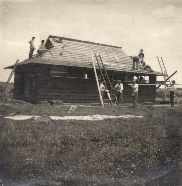 A group of men constructing a bungalow at University Settlement Camp. Three ladders are being used and five men are on the roof.