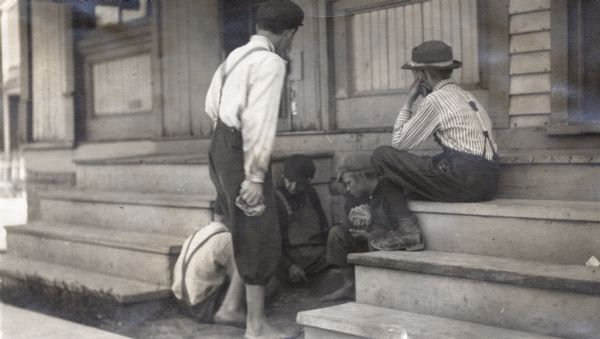 A group of boys play a game of cards while sitting on the ground between the steps of some houses. At least four boys wear caps, three are wearing pants with suspenders, and two are wearing overalls.