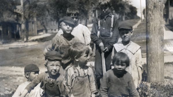 A portrait of eight young boys posed standing and sitting on the side of a street. Four of the boys wear caps, and along the street in the background are houses.