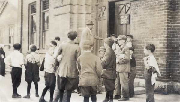 A group of boys are standing near the entrance of a brick building waiting to get in. A man behind them is entering the door. The sign near the door reads "Gents Day."