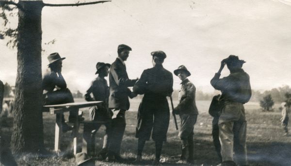 Boy Scout Leaders having a meeting while on a hike. They are all standing near a tree in a field, and one man is seated on a bench near the tree.