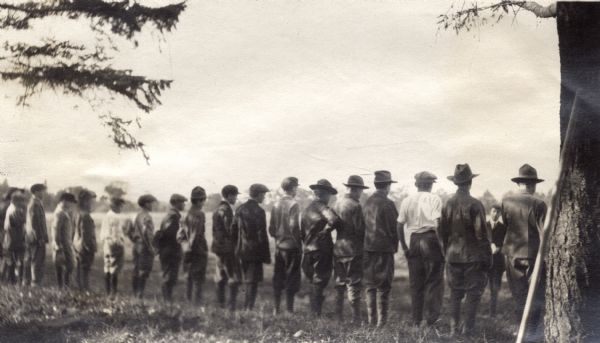 Rear view of group of boy scouts standing at attention in a line in a field near some trees. One man stands in the middle addressing the group.