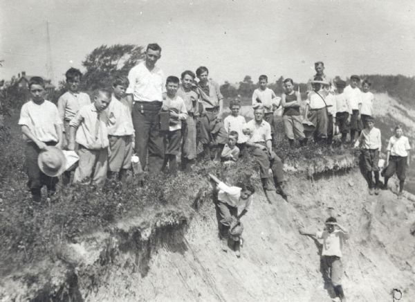 A Y.M.C.A. hiking group of men and boys posed standing on the edge of a sandy drop in a field. A house and trees are in the background. Most of the group is wearing dark-colored pants and a light-colored shirt, and some have hats.