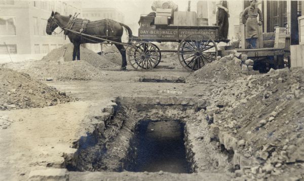 Buffalo Street construction showing cuts in the pavement and the inconvenience for traffic. Standing between the excavated holes and piles of dirt is a horse pulling a heavily-loaded horse-drawn wagon, with two men in work clothes standing near a wood platform in front of a storefront or commercial building.