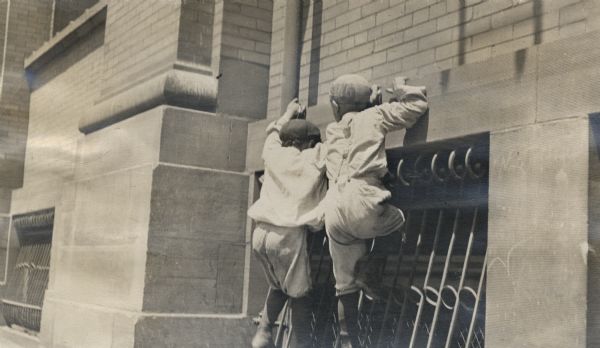 Two boys attempting to climb a wall on the facade of a building near a window.