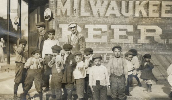 A group of boys take a boxing stance near two men standing on a street corner, in front of a building that has "Milwaukee Beer" painted on it.