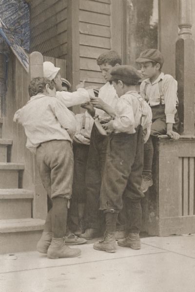 A group of young boys gathered at the steps at the front of a house engaging in some kind of game, perhaps a game of chance. One boy is sitting above them on a railing.