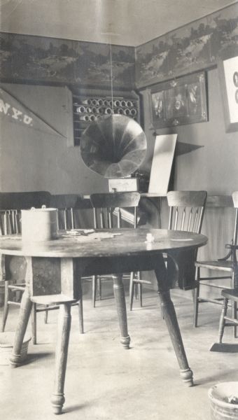 Interior of a clubhouse with a table, chairs, wall decoration, and a phonograph. Possibly the Can-Crushing Club.