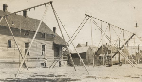 A deserted playground with swing sets, slides, and ladders, in front of a group of buildings. There are two young boys near a dwelling in the background.