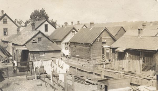 Elevated view of backyard areas, including some dilapidated houses and outbuildings. There are wood fences surrounding some of the yards, and laundry hangs from lines in the foreground.