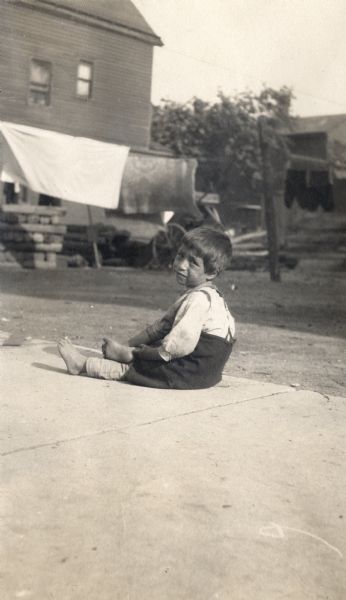 A barefoot child is sitting on the sidewalk. Houses and buildings are in the background, and laundry is hanging out to dry.