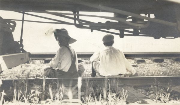 Two boys sitting on the railroad tracks under a train. The boy on the left is looking back over his shoulder and is wearing a straw hat with a small American flag attached to it.