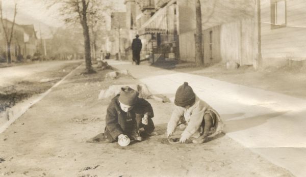 Two children making mud pies in the dirt in the space between the street and sidewalk. They are dressed in warm clothes and hats. In the background a man is walking down the sidewalk near an awning, perhaps a storefront.