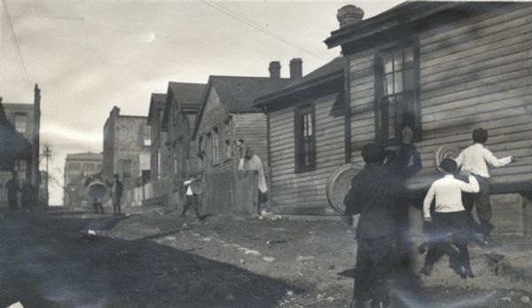 Children having a stone fight in an unpaved alley or street in an underprivileged neighborhood. Some of the boys are holding trash can lids as protection, and a woman is leaning on a fence watching them.