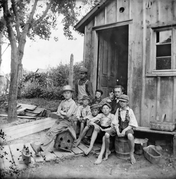 Small group of boys and one man sit outside the open doorway of a small wooden house or shed.