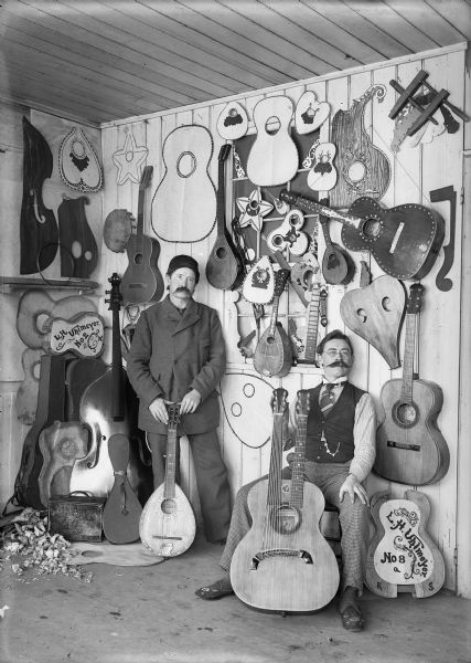 Two men posing with guitars, one seated and one standing. They are probably in a music store or a musical instrument manufacturer. Several guitars and mandolins hang on the wall.