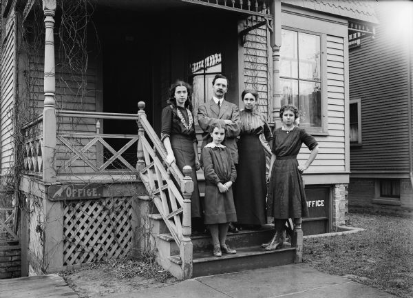 Father, mother and three girls stand on front porch of what appears to be their home. There is a sign for an office in the basement on the left.