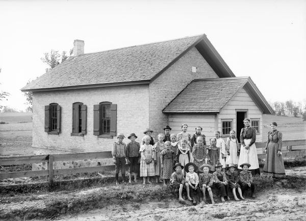 Large group of children and two women standing in neat rows with two women outside of fence in front of a brick building that appears to be a country school house.