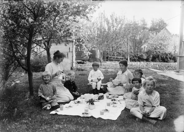 Six children and two women sitting on the lawn with picnic food and beverages. There appears to be a garden in the background.