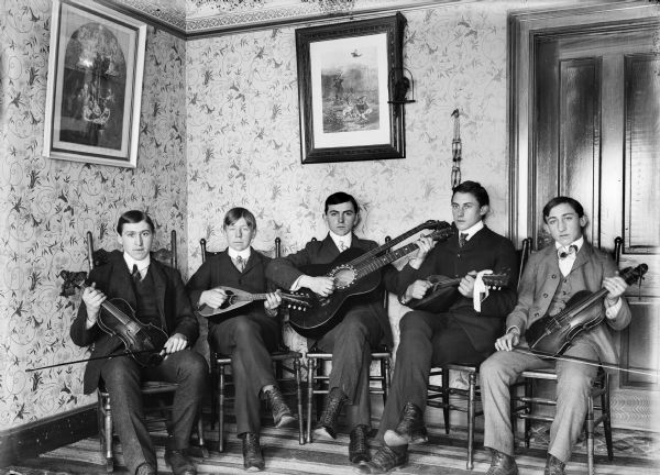 Group of five men sitting and holding stringed instruments. Two of the men hold violins, two hold mandolins, and one man holds a guitar.