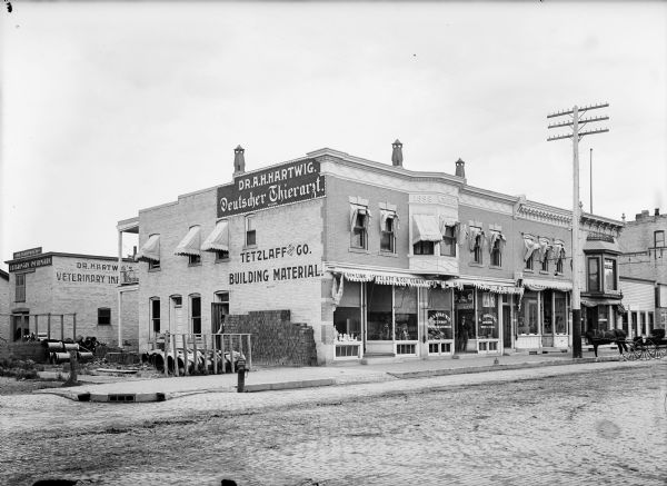 View from Third and Main Street of storefronts. There is signage for the Tetzlaff and Co. Building Materials above a pile of bricks in a sideyard near the storefront. Next door is the Hartwig Veterinary storefront. The Hartwig Veterinary Infirmary building is in the background on the left.