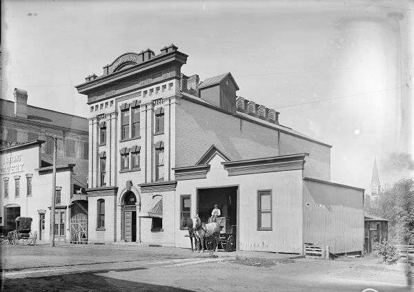 Jacob Jossi building with man sitting on horse-drawn wagon in open doorway. A buggy is parked in front of G.W. Evans Livery. The steeple of a church building is in the background.