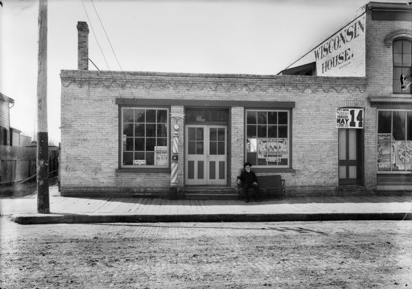 View from road of barbershop storefront in Watertown. The sign on top of the barbershop pole says: "L. Spangenberg." A man wearing a hat sits on a bench in front of the shop on the wooden sidewalk. A building next door has a sign painted on the side for the "Wisconsin House." Other signs advertise the "Seibel Bros. World's Greatest Dog and Pony show."