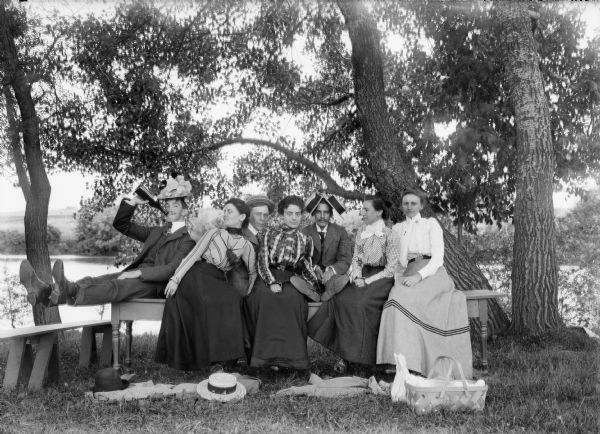 Group of three men in comical poses sitting with four women on a bench in wooded area with a lake behind them.