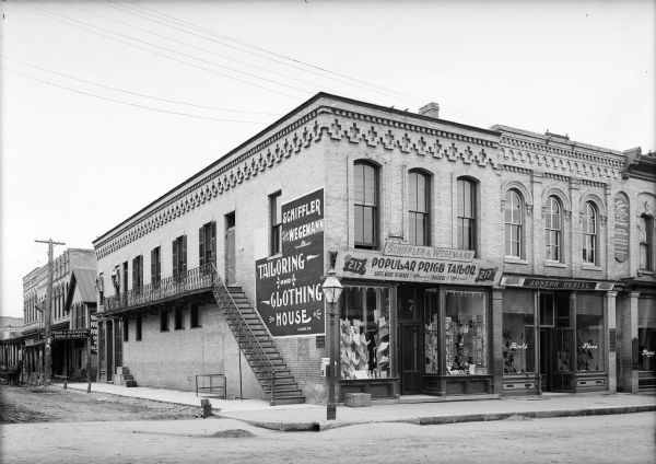 View from road of Schiffler and Wegemann Tailoring and Clothing House building on corner. The storefront is at Third and Main Streets. On the left side of the building is a flight of steps to a second floor balcony. Next door on the right is Joseph Hertel Boots and Shoes.