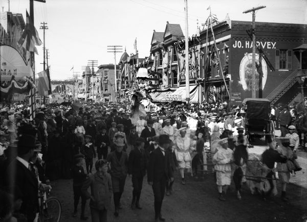 Parade scene with a woman on a float, and costumed children and horses. The storefronts and street are decorated with flags and bunting. An archway has been set up over the street in the background.