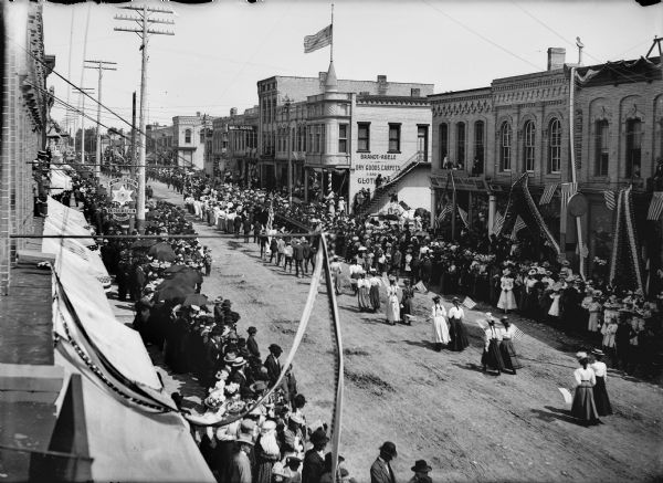 Elevated view of parade. Crowds are gathered on either side of the street, many holding umbrellas. Women are walking two-by-two carrying flags in parade. Flags and bunting decorate storefronts.