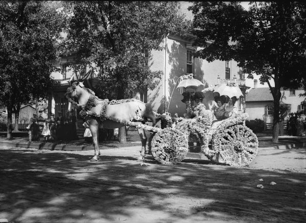 Horse-drawn carriage elaborately decorated with flowers appears to be stopped off parade route. The carriage is carrying four women wearing formal dresses and fancy hats and holding umbrellas.