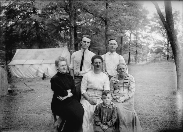 Five adults and one child pose outside in what appears to be a park, with two tents in the background.