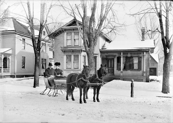 Winter scene of couple in horse-drawn sleigh on residential street.