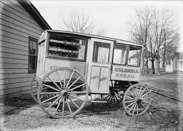 Side view of the Columbia Bread Cart with the address written on it: "618 Main St." There is a building on the left, and in the background are other buildings and trees.