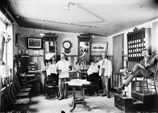Interior of barbershop. Two barbers stand by barber chairs, and one younger man sits on shoe shine stand on the right.