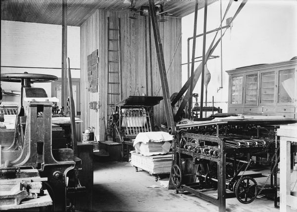 Interior of print shop. The machinery, including printing presses, are belt-driven. The belts are driven by machines attached to the top of the high ceiling.