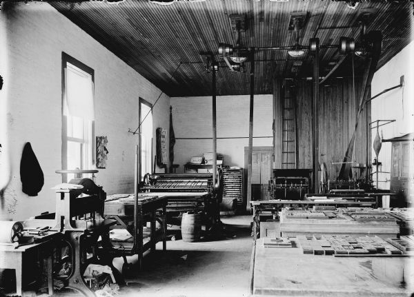Printing presses are towards the back of the long, narrow room, and in the foreground are tables with type laid out in form or frame for letterpress printing. The machinery in the print shop is belt-driven. The belts are driven by machines attached to the top of the high ceiling.